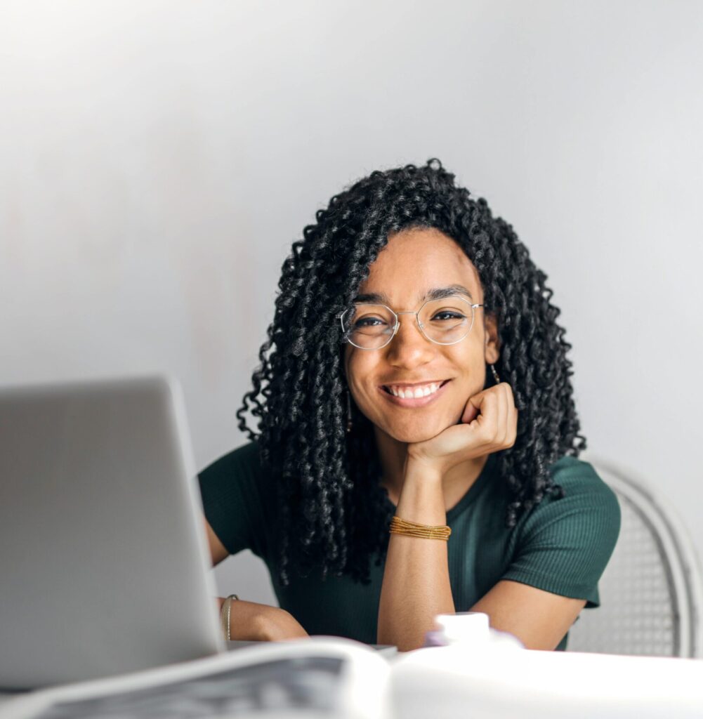 Joyful businesswoman with curly hair smiling at camera while using laptop indoors.